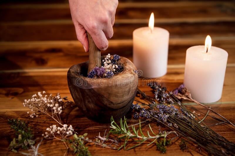 A hand grinding dried herbs with a mortar and pestle, flanked by lit white candles, depicting a ritual for Candle Healing & Spell Casting.