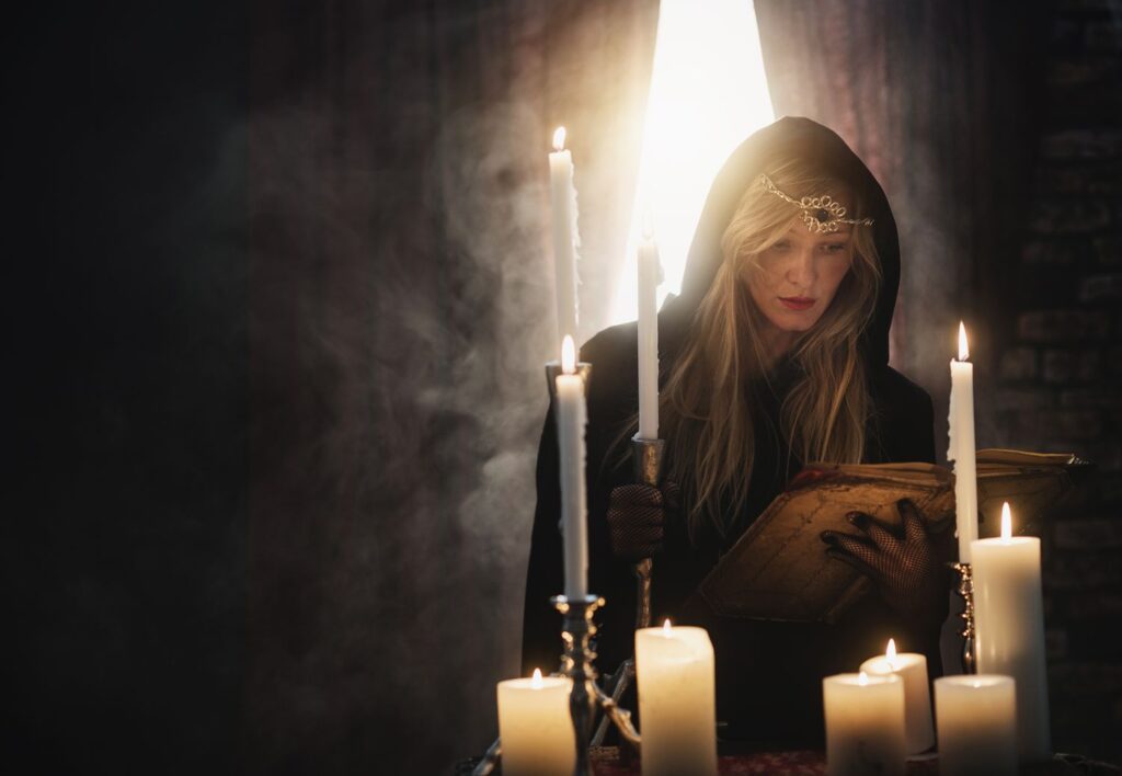 A mysterious woman in a hooded robe reading from an ancient book, surrounded by tall candles, illustrating the art of Candle Healing & Spell Casting.